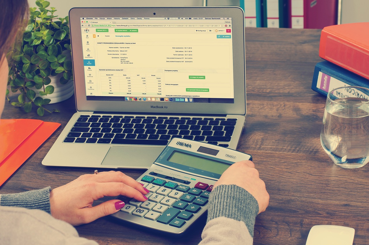 Une personne utilise une calculatrice tout en travaillant sur un document financier affiché sur un ordinateur portable avec Chorus Pro ouvert. L'espace de travail comprend une plante en pot, un verre d'eau et un dossier contenant des papiers, le tout sur une table en bois. La scène suggère un travail de bureau ou une planification financière.